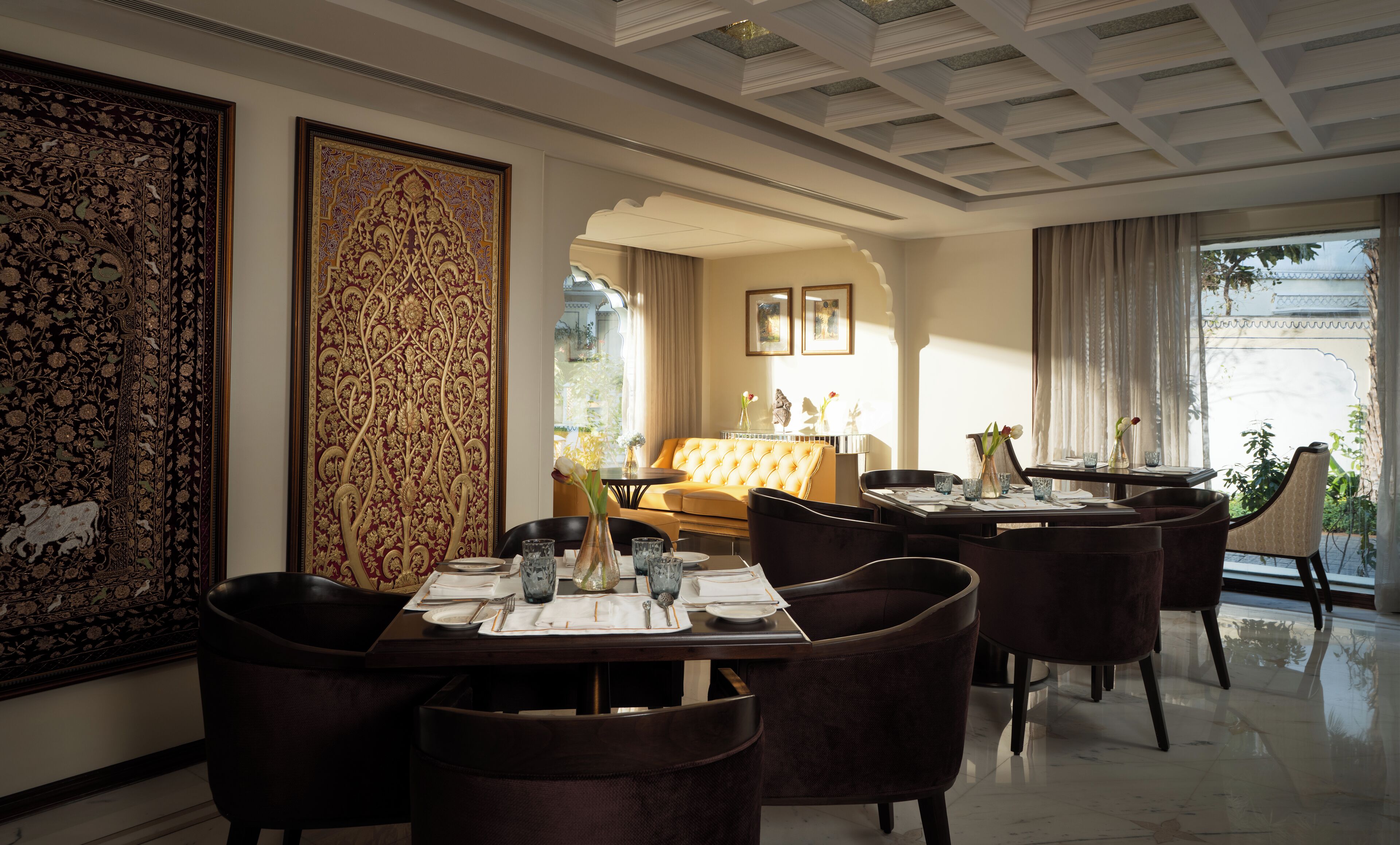 A dining area with a table set for four, surrounded by dark upholstered chairs. A large, ornate tapestry hangs on the wall to the left.