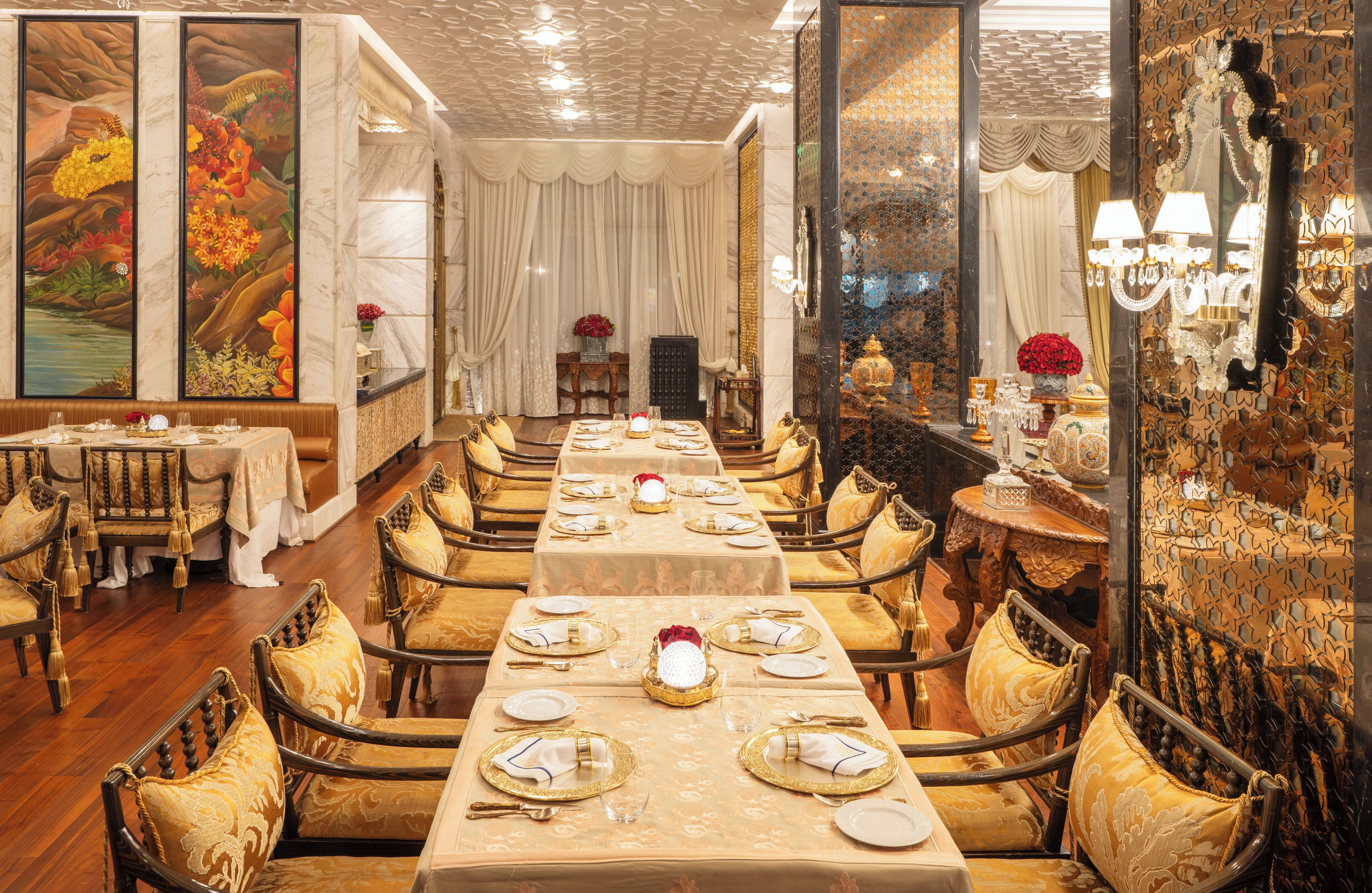 A long dining table set with gold accents, surrounded by ornate chairs, in a room with patterned walls and a chandelier.