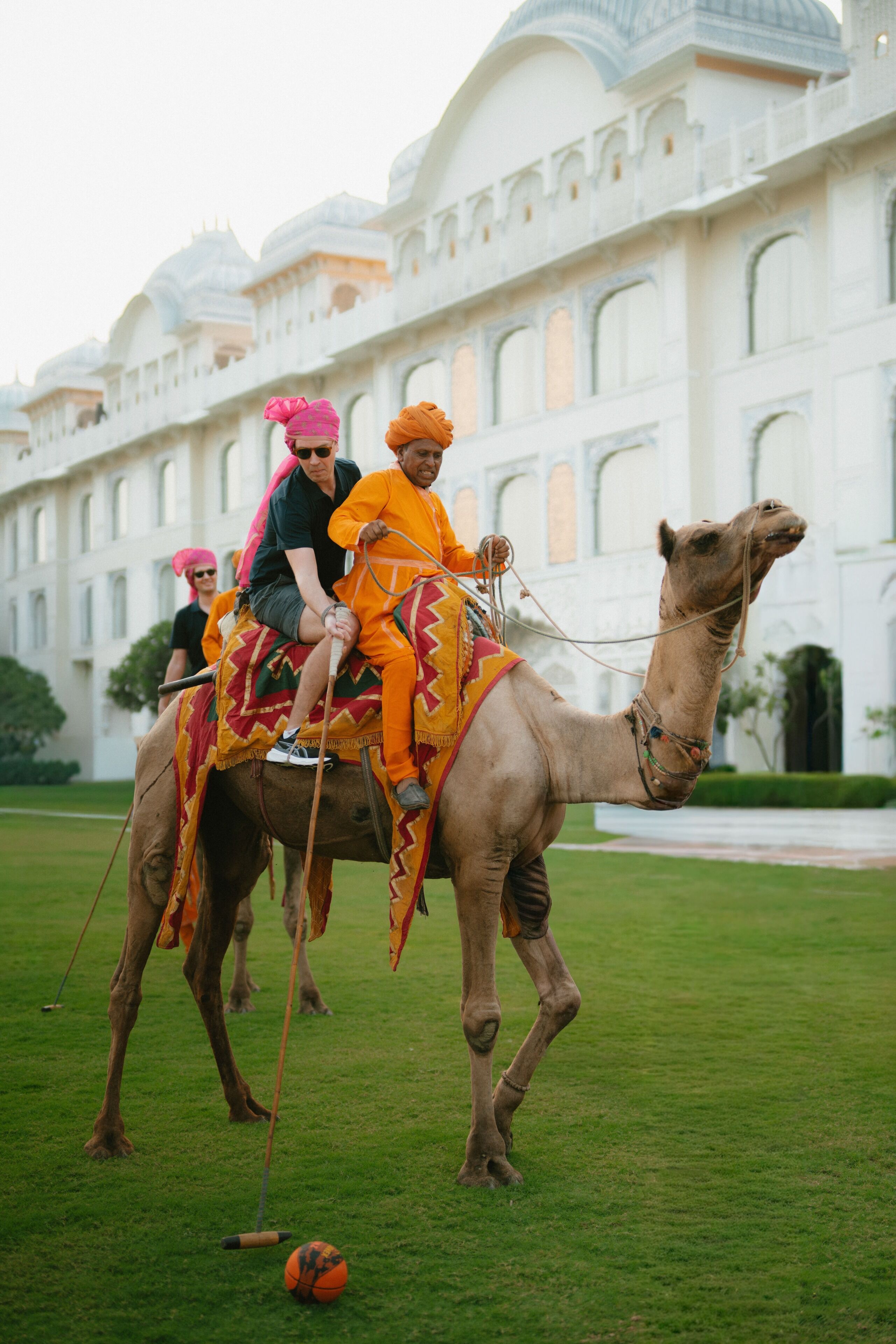 A camel with riders in traditional attire, a grassy area, and a large white building with arches in the background.