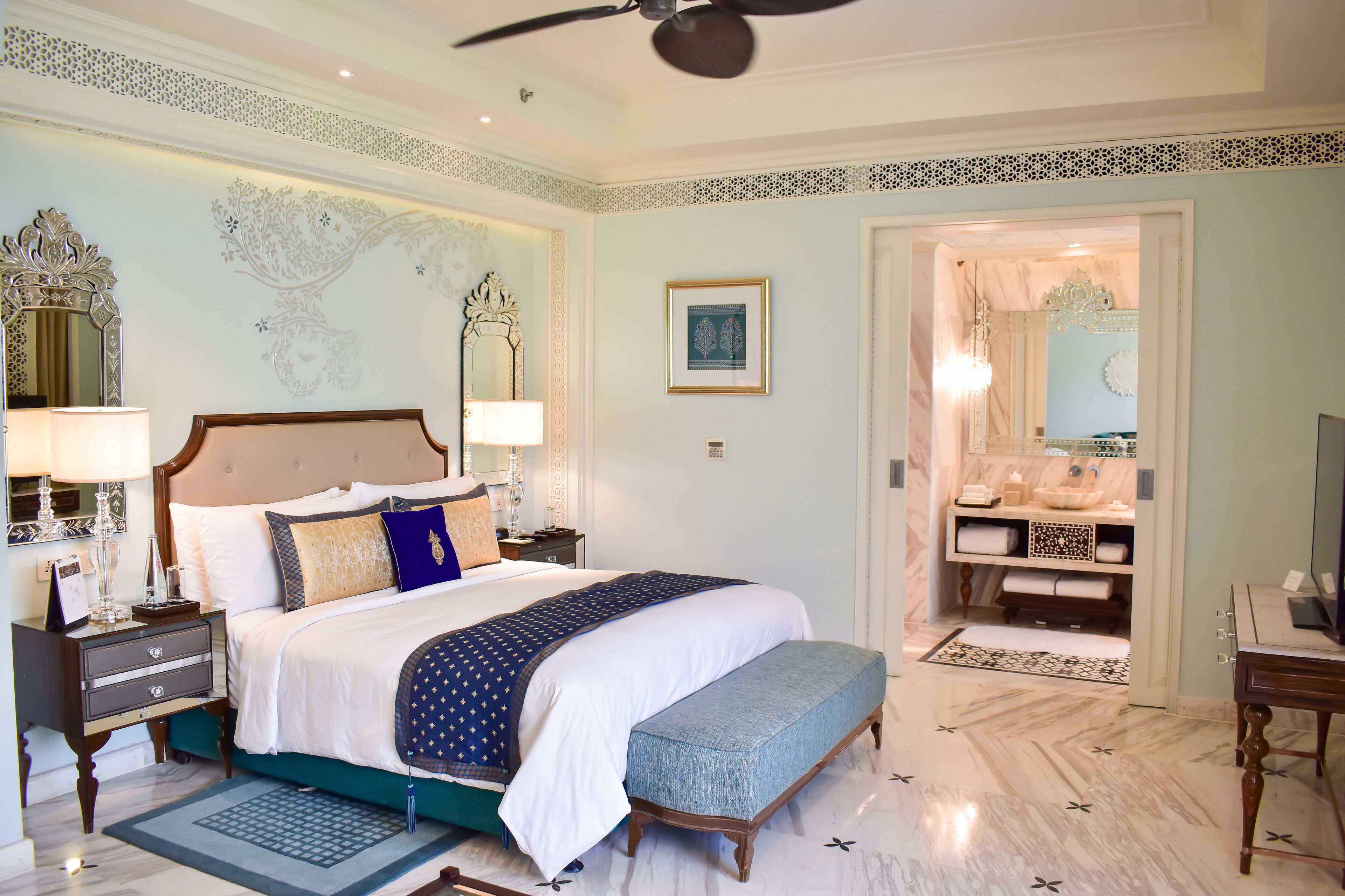 A spacious bedroom with a large bed, a sitting bench, a vanity with a mirror, and a television.