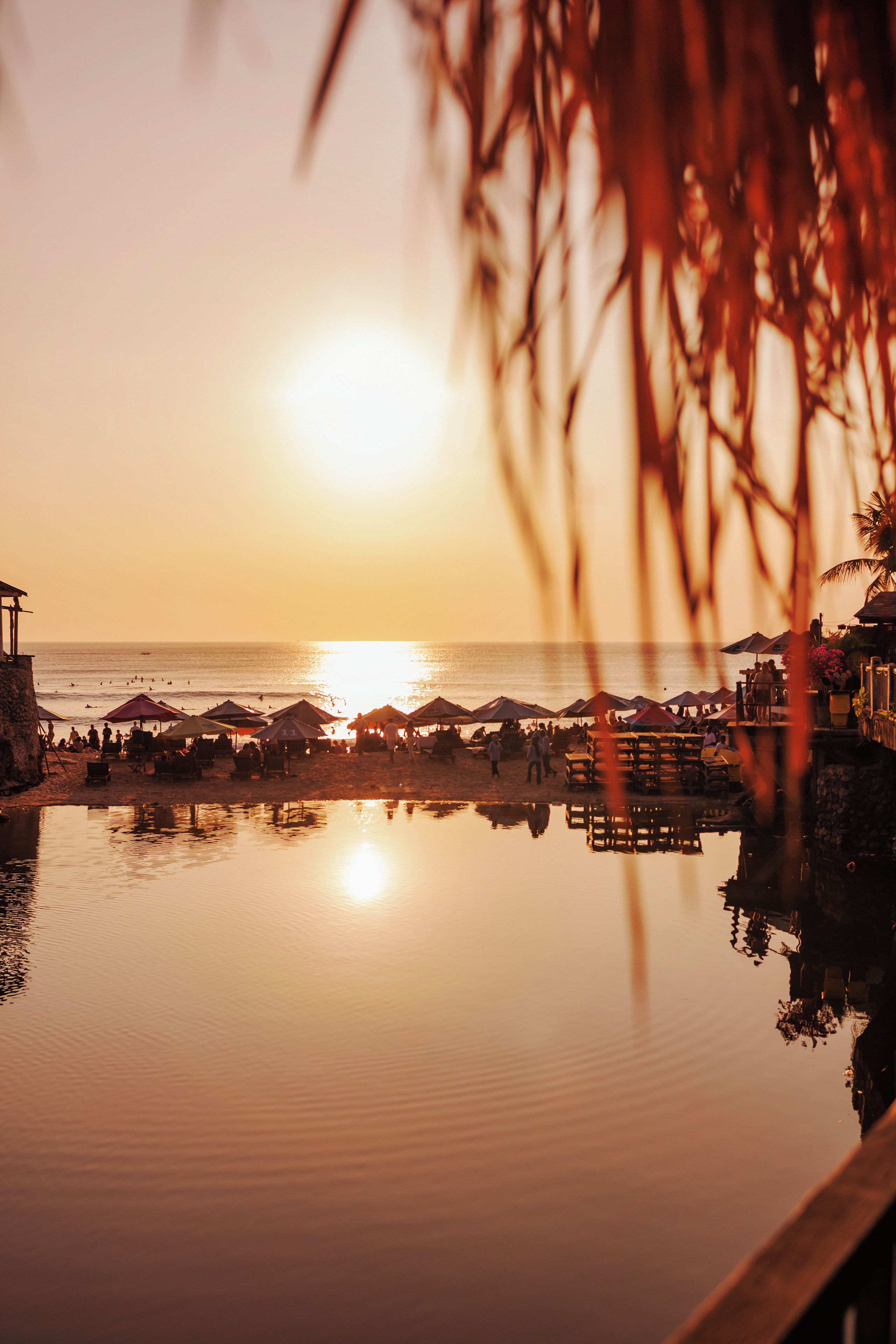 Une scène de plage avec une étendue d’eau calme reflétant le coucher de soleil, des parasols et quelques personnes.