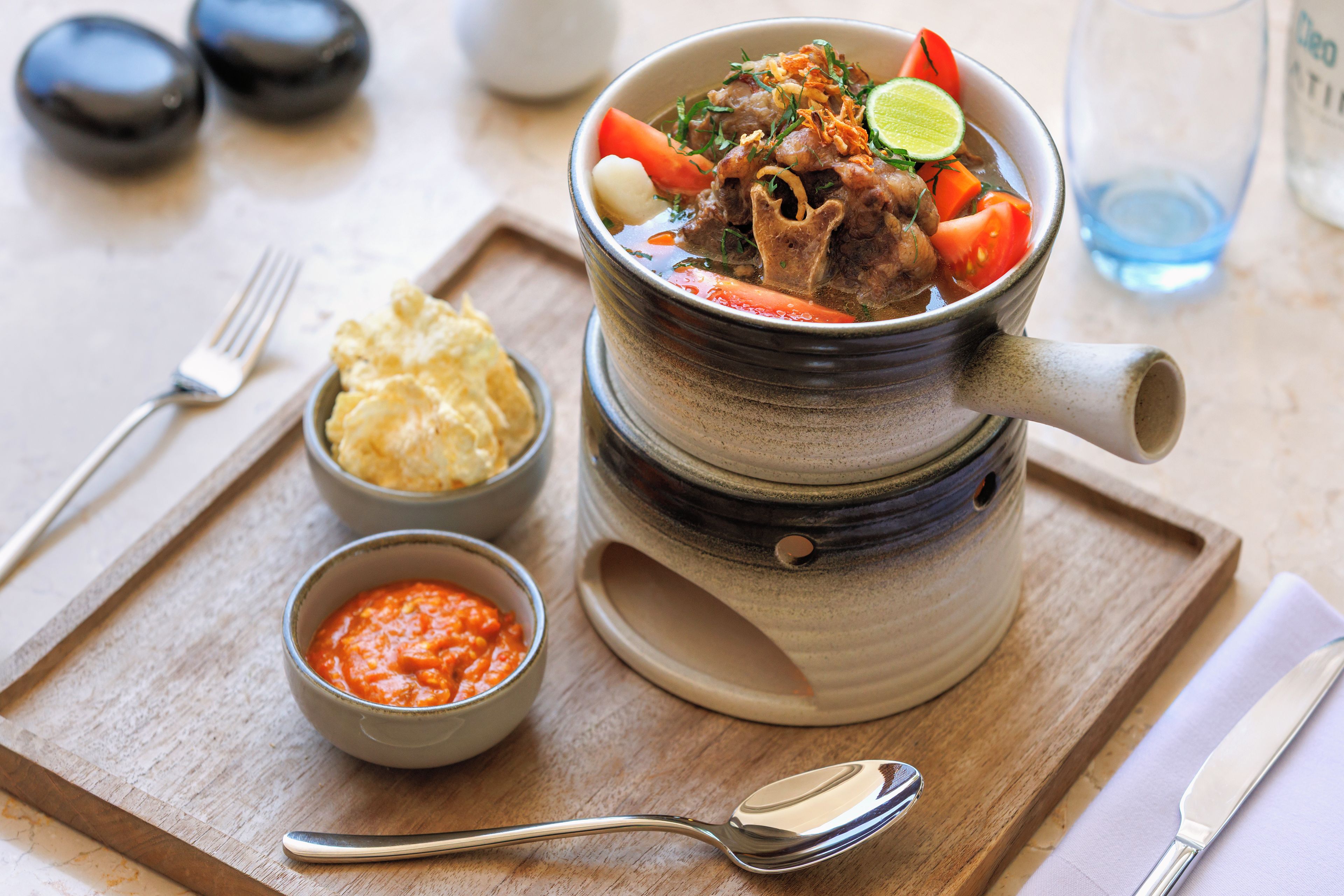 A meal served in a traditional clay pot with a spoon and fork on a wooden tray.