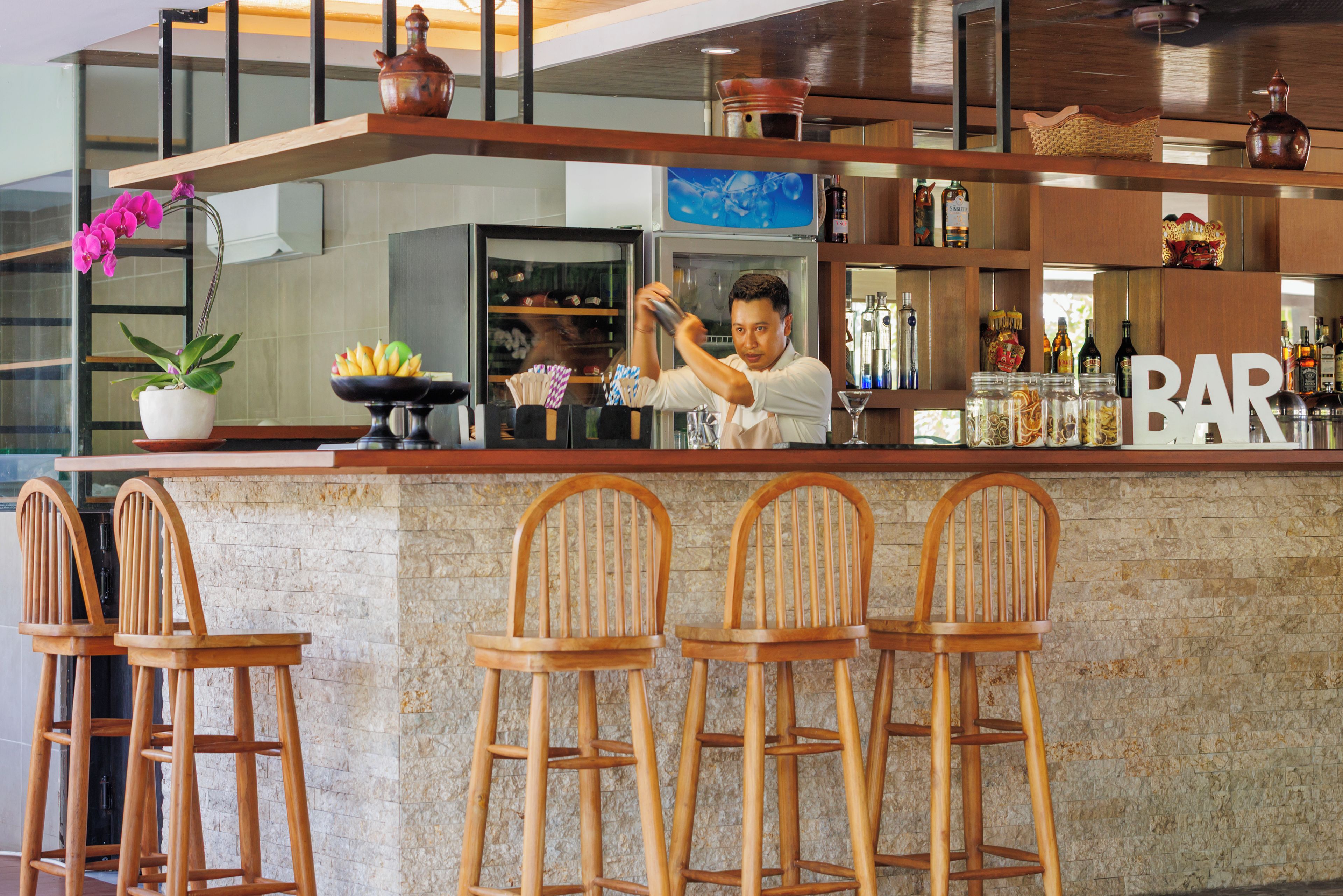 Zona de bar con un cantinero, taburetes de madera y una variedad de botellas y vasos.