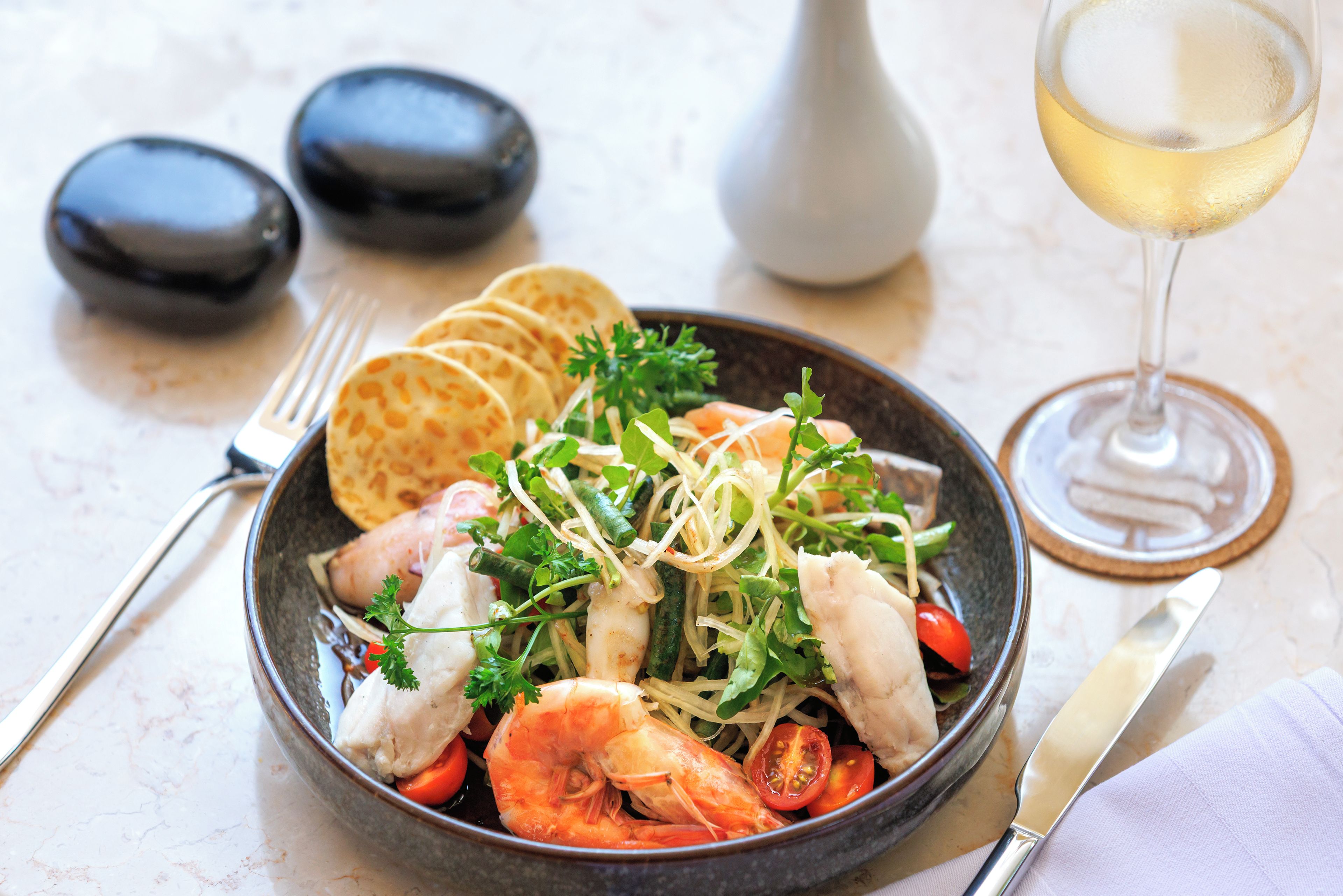 A bowl of seafood salad with shrimp, vegetables, and herbs, accompanied by a glass of white wine and a fork on a marble table.