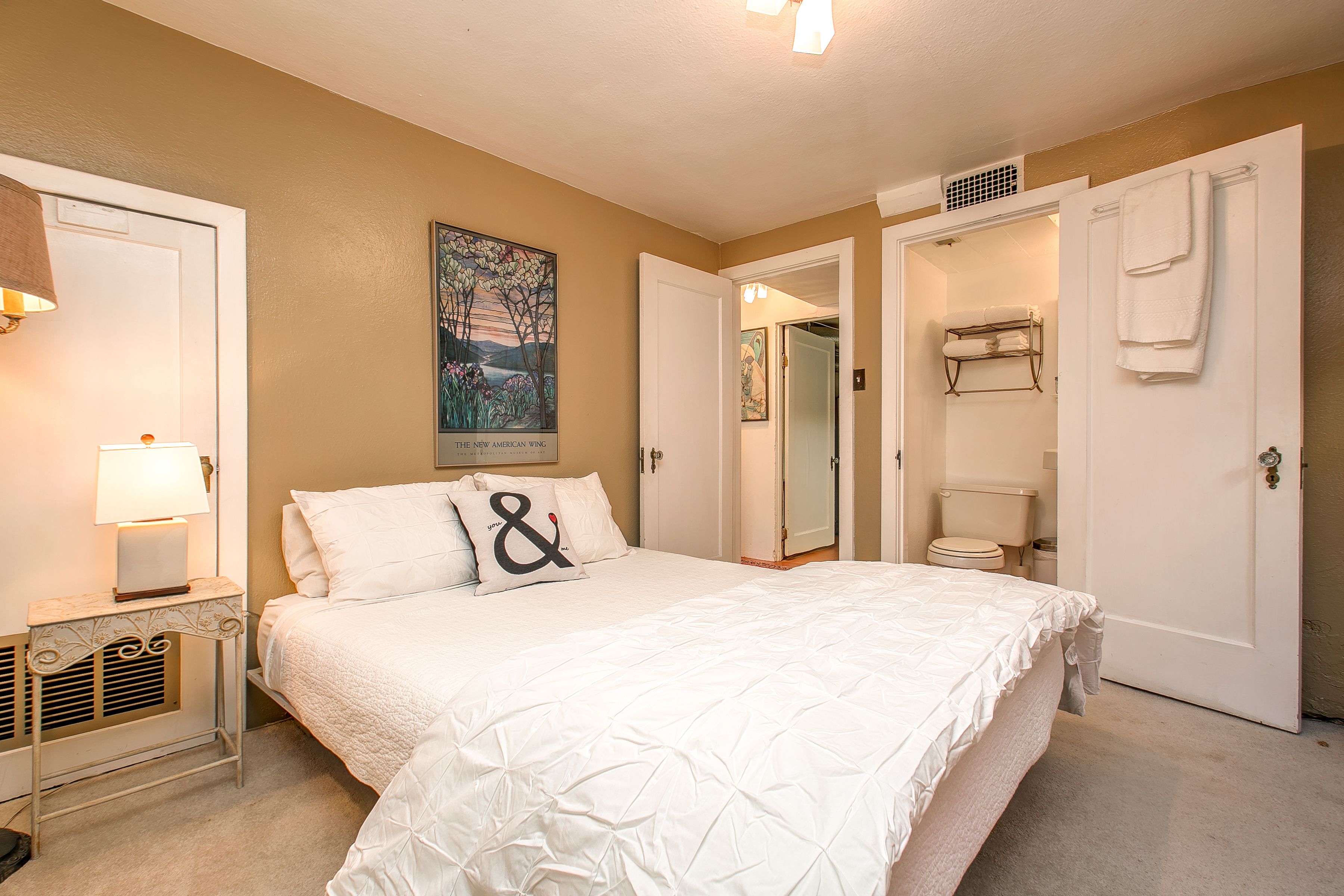 A bedroom with a bed, a nightstand, a painting, a bathroom with a toilet, and towels on a rack.