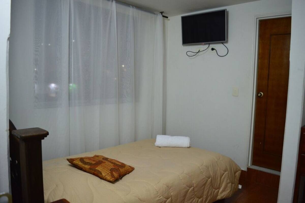 A bedroom with a bed, a TV mounted on the wall, a wooden door, and sheer curtains.