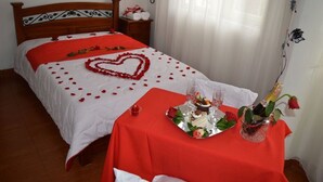 A neatly made bed with a heart-shaped decoration and rose petals, a small table with a tray of desserts and champagne, and a bedside table with towels and a phone. - Hotel Dancar (Pamplona)