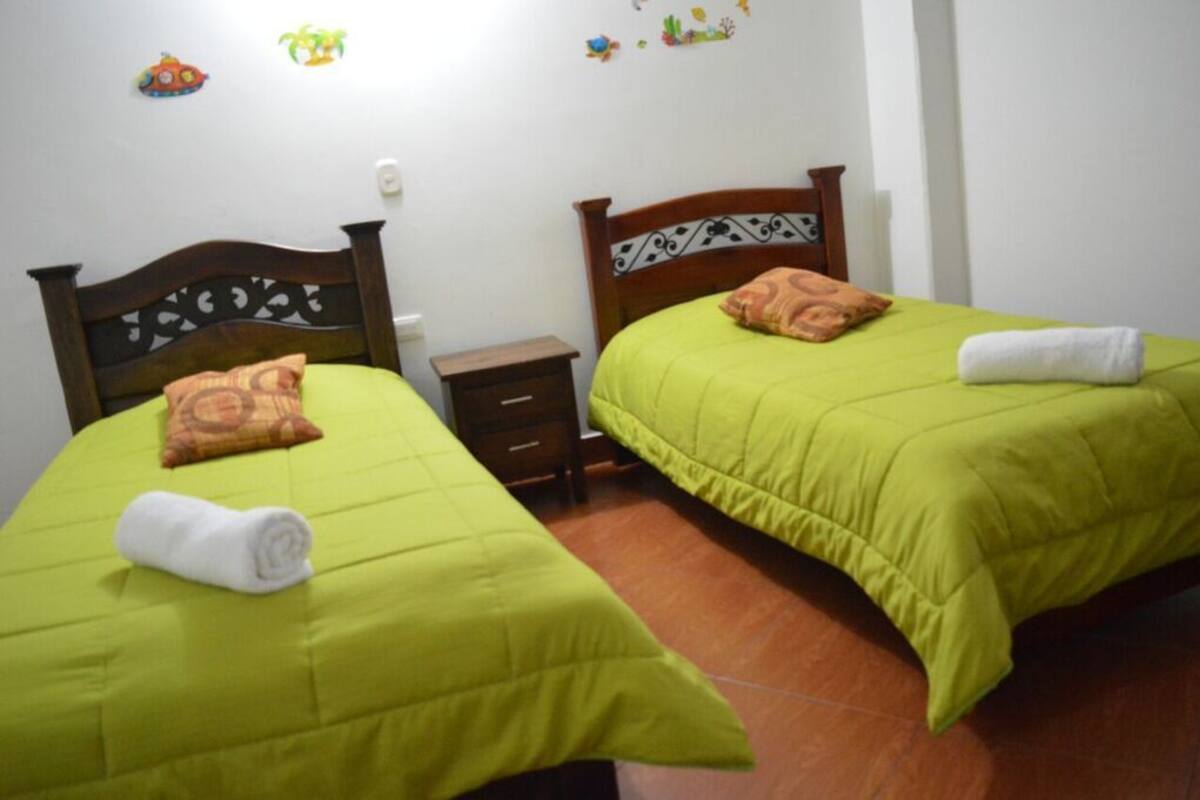 A room with two beds, each with a green bedspread and a rolled white towel on top. There is a small wooden nightstand between the beds.