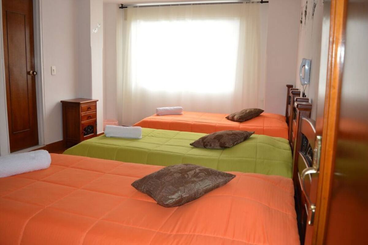 A hotel room with two beds, each with a different colored bedspread, a wooden wardrobe, a small table with a lamp, and a window with curtains.