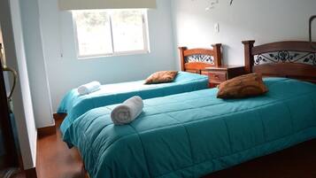 Una habitación con dos camas, cada una con ropa de cama color turquesa y almohadas marrones, un cabecero de madera, una mesita de noche pequeña y una ventana con vistas.