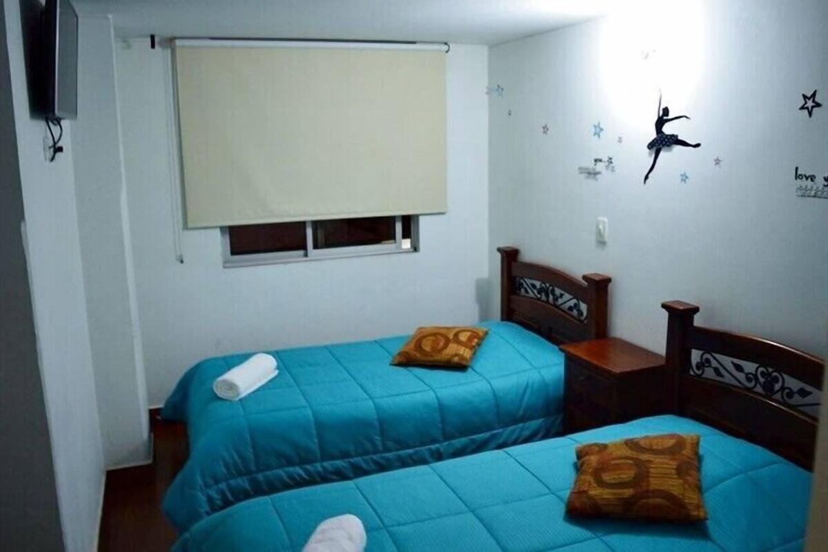 A room with two beds, each with a blue bedspread and a brown pillow. There is a window with a beige blind, a wall-mounted TV, and decorative star stickers.
