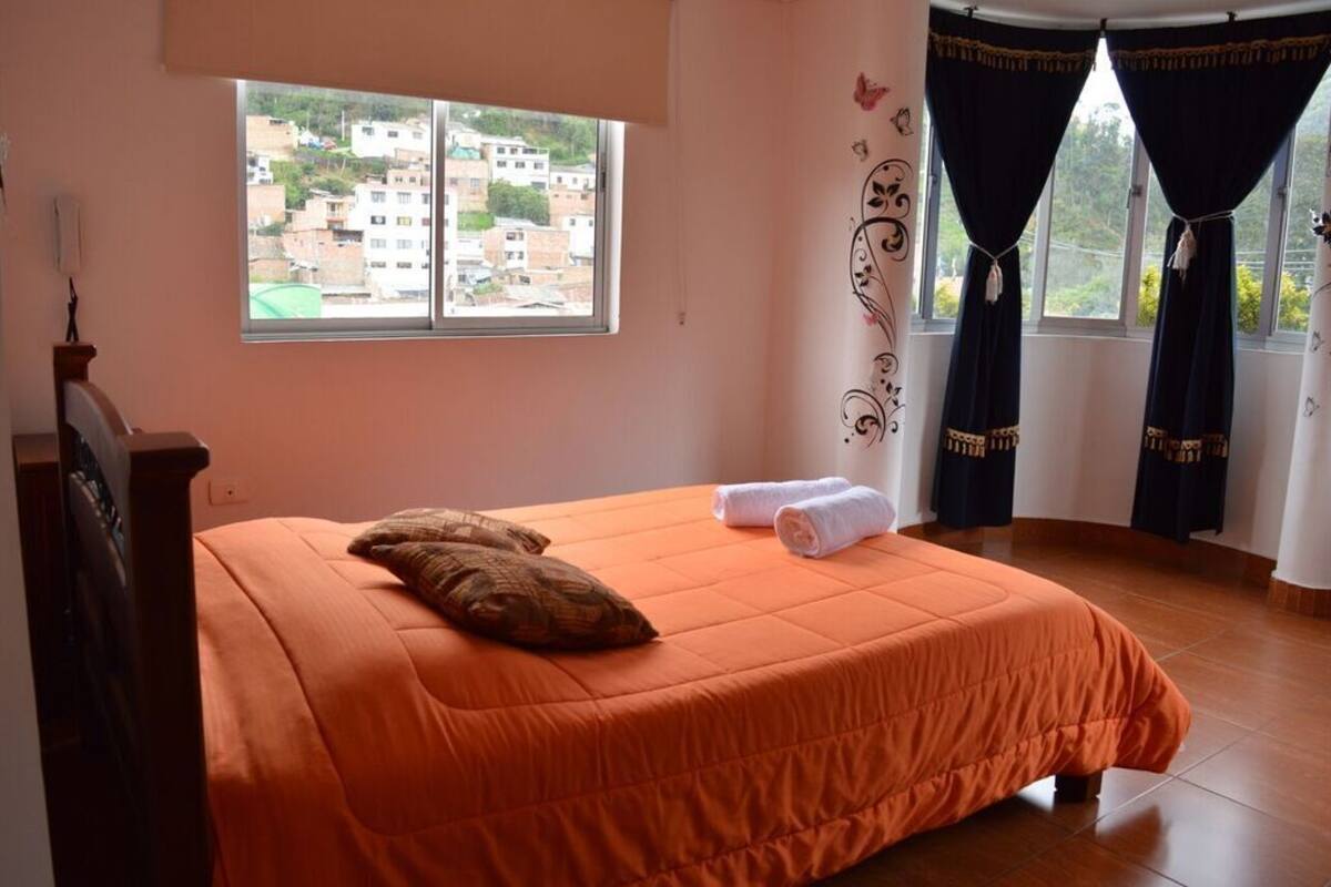 A bedroom with a bed, a window, curtains, and a view of buildings.