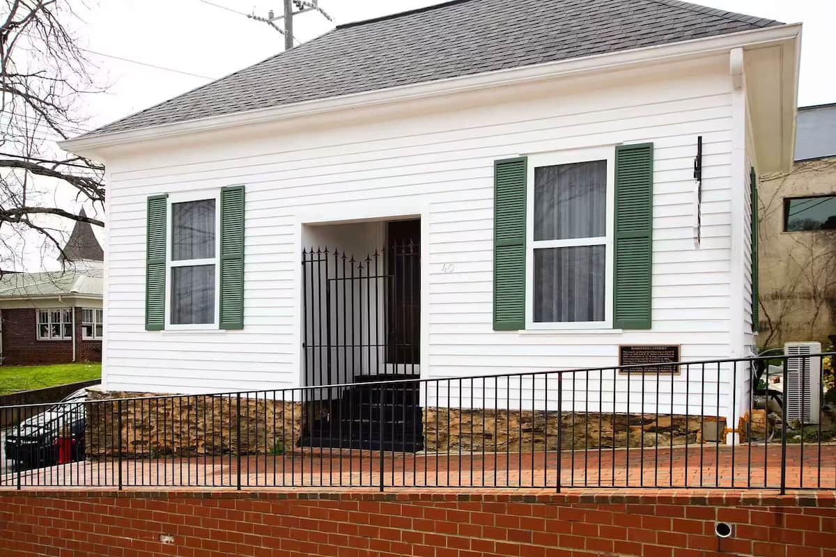 A white building with green shutters, a black door, and a brick foundation.