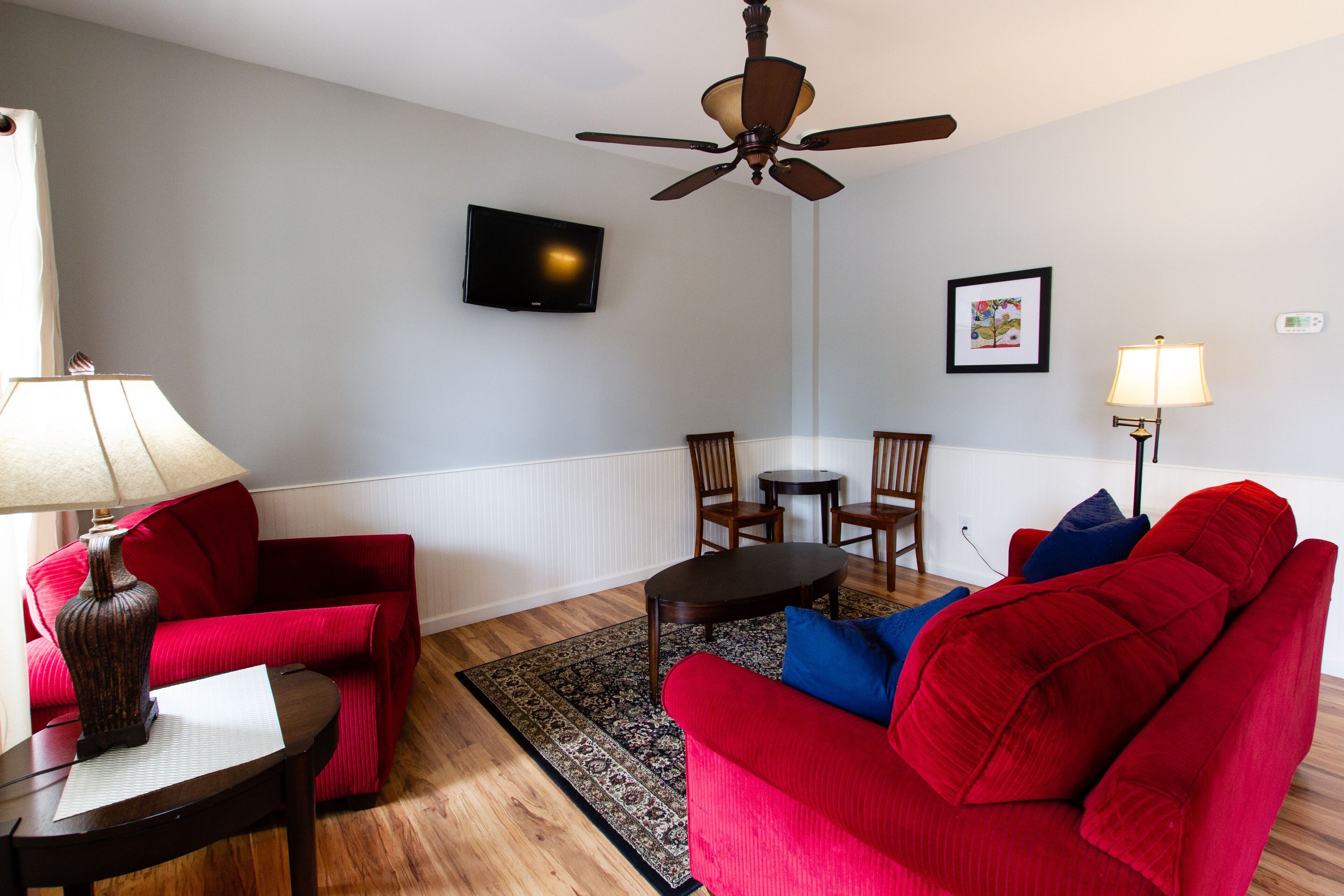 A living room with a red sofa, a red armchair, a wooden coffee table, a floor lamp, a ceiling fan, a flat-screen TV, and a framed picture on the wall.