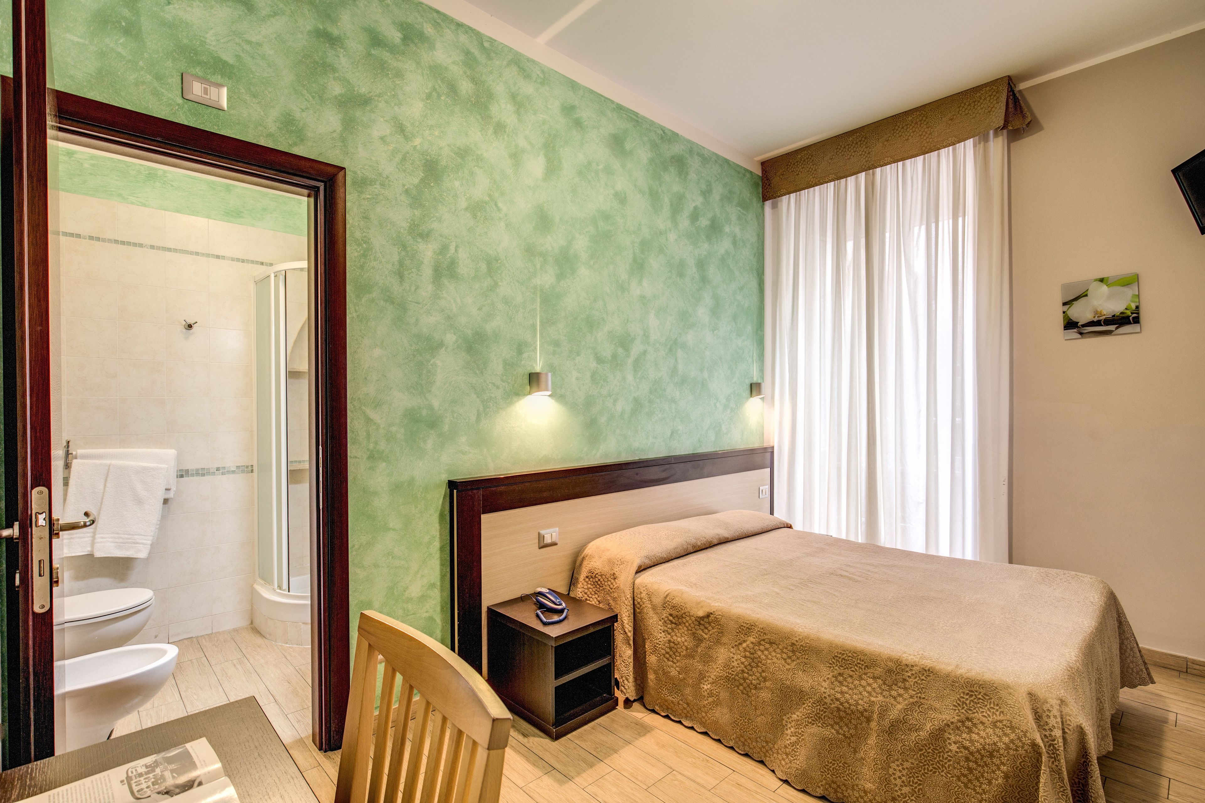 A hotel room with a green wall, a wooden headboard, a bed with a beige blanket, a nightstand with a phone, and a view of a bathroom with a toilet and shower.