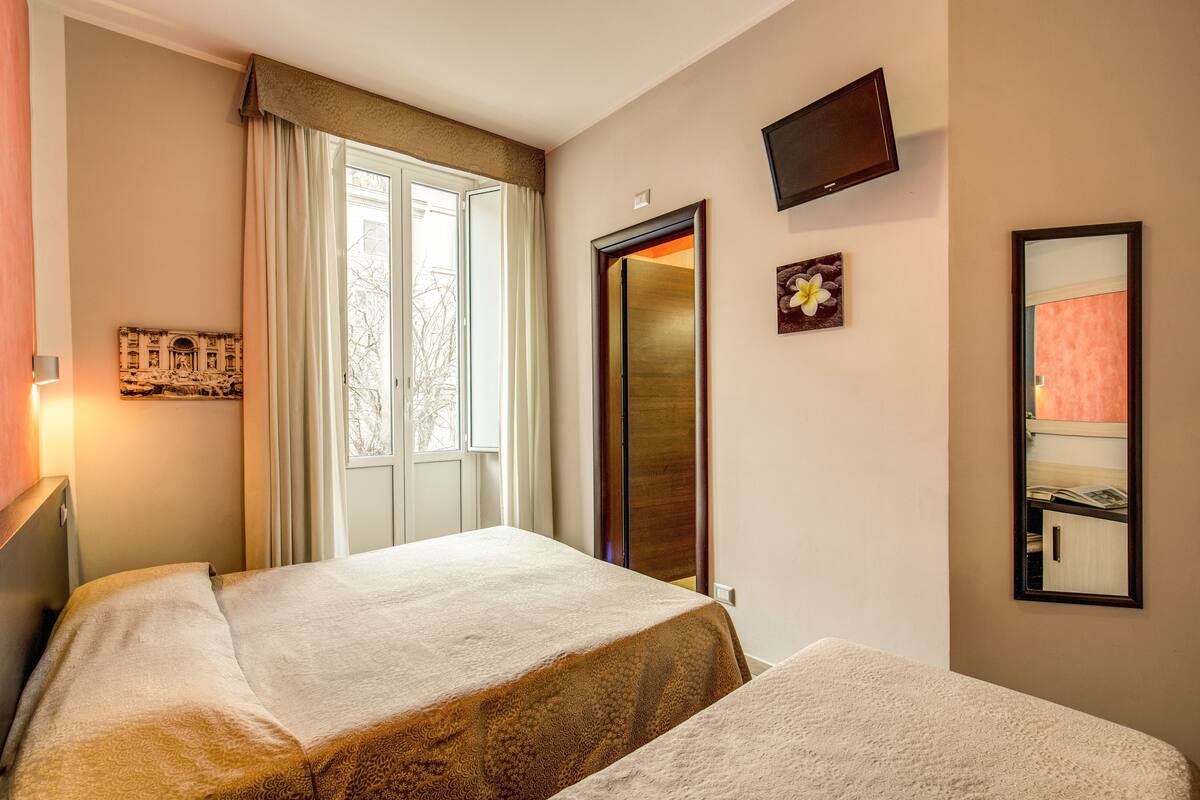 A hotel room with two beds, a large window with curtains, a flat-screen TV, and a mirror on the wall.