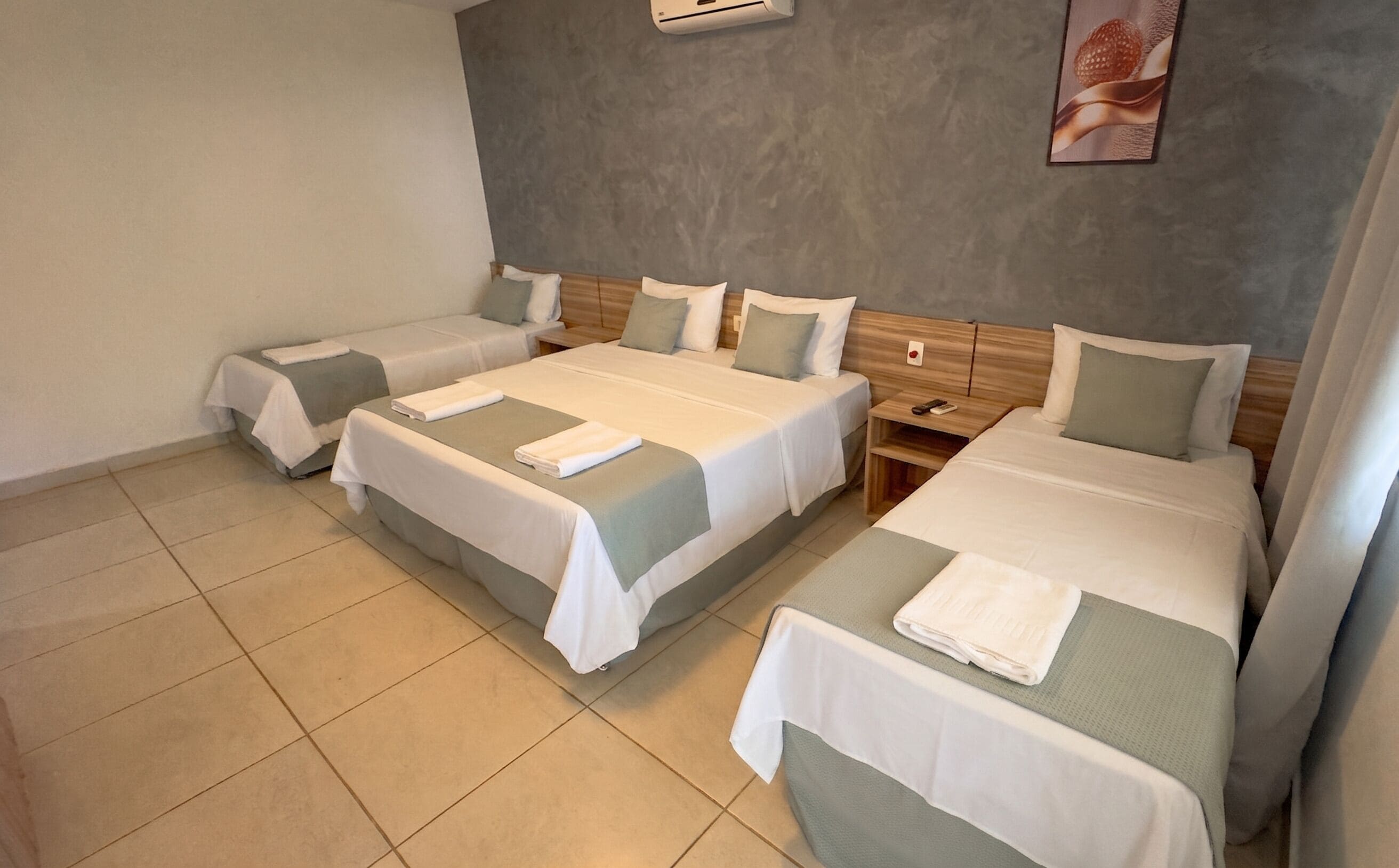 Una habitación de hotel con tres camas, una pared gris con textura y suelo de baldosas.
