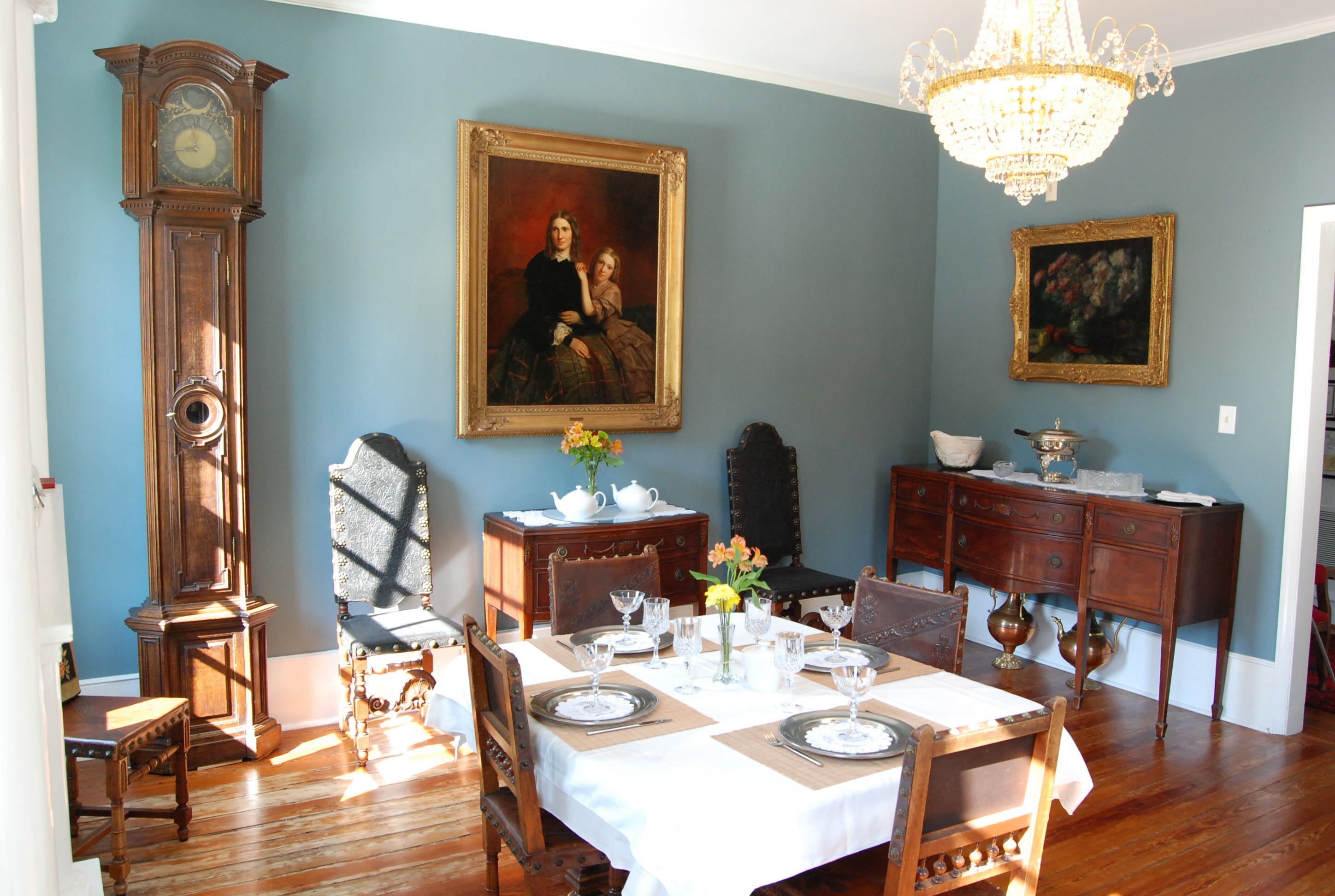 A dining room with a wooden table set for four, surrounded by antique chairs, a grandfather clock, and two paintings on the wall.