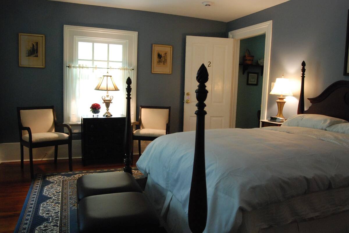 A bedroom with a four-poster bed, two chairs, a desk with a lamp, and a door with the number 2.