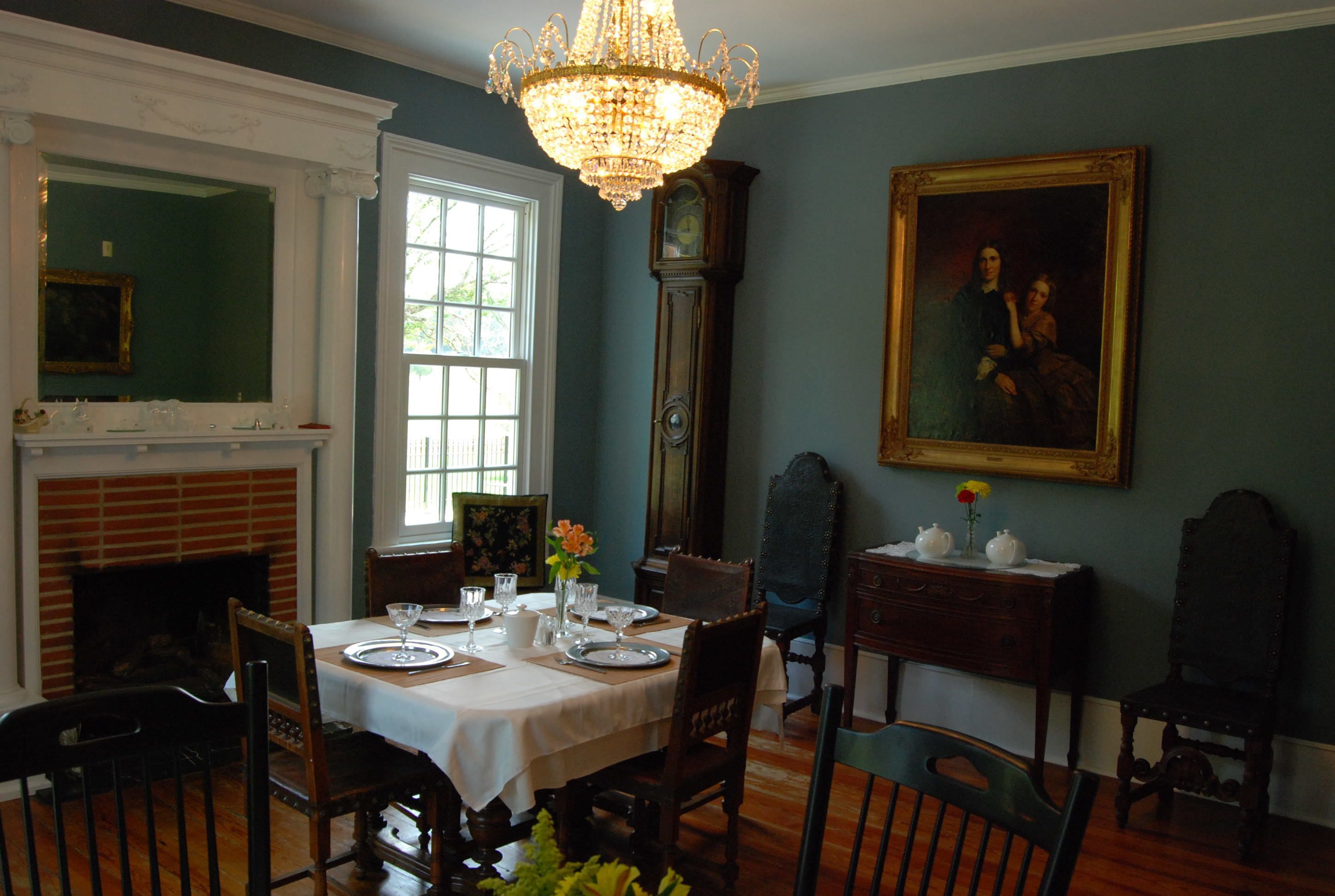 A dining room with a table set for four, a grandfather clock, a fireplace, and a framed portrait on the wall.