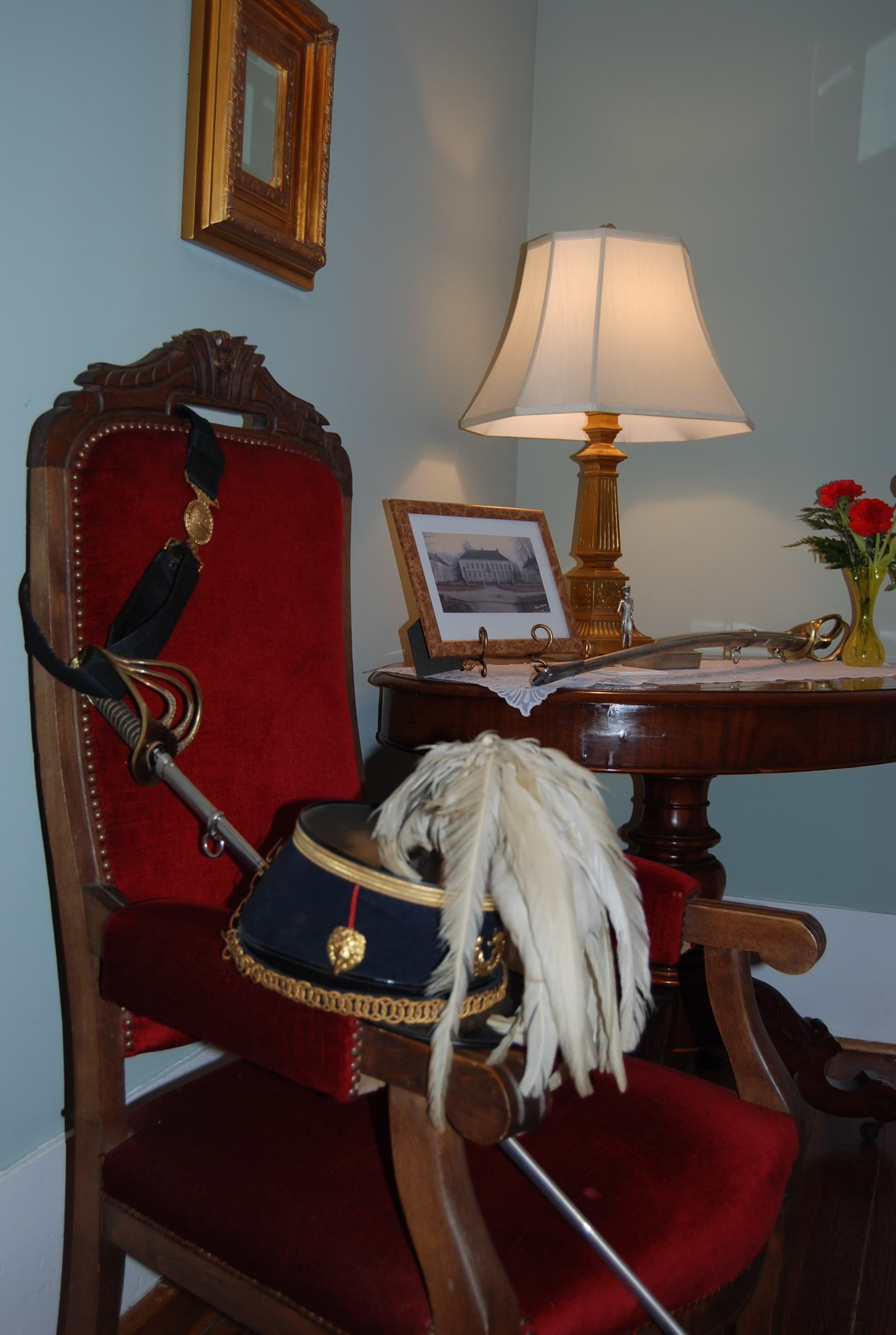 Una habitación con una silla tapizada de rojo, una mesa de madera con una lámpara, un cuadro enmarco y un florero con flores.