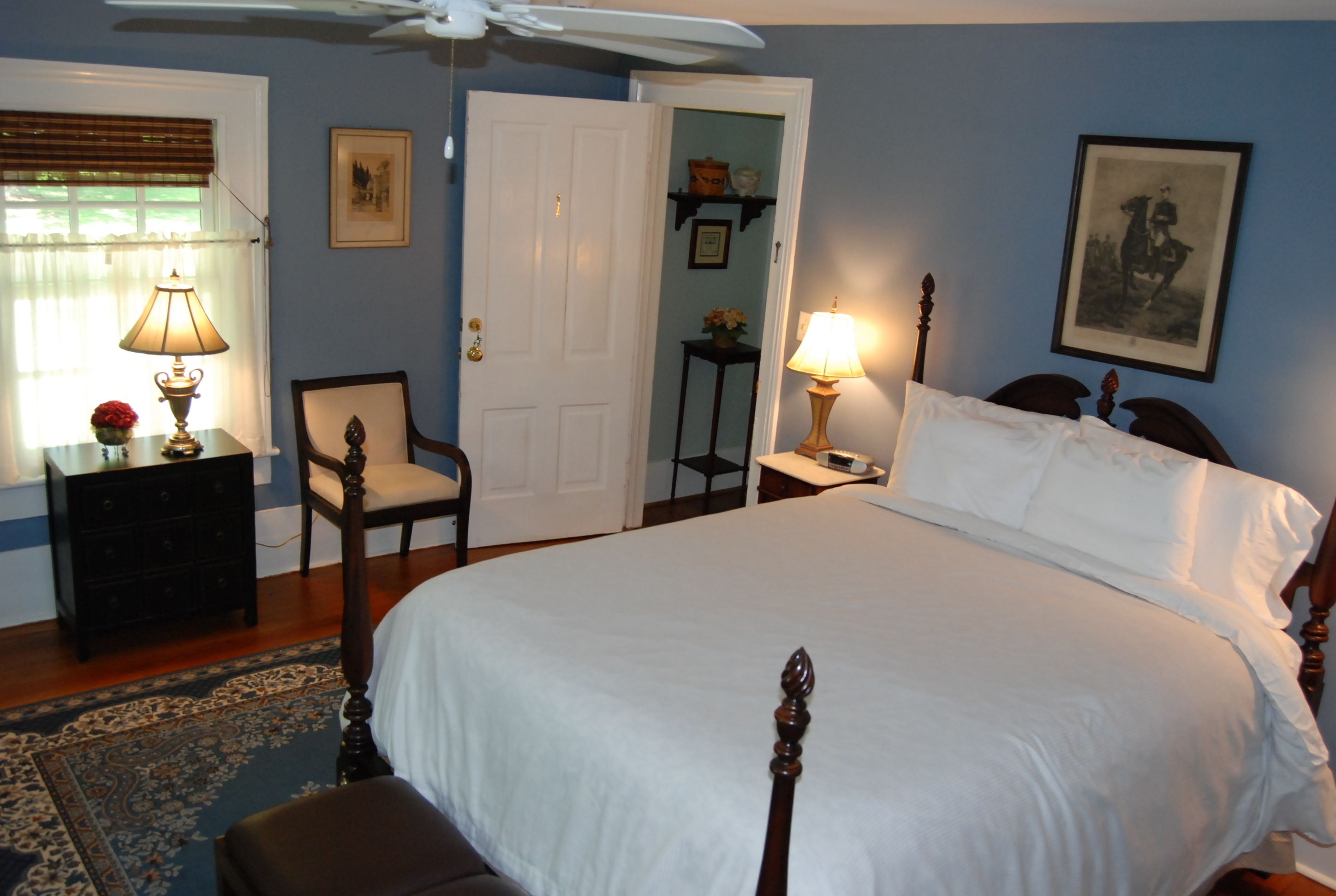Une chambre à coucher avec un grand lit, deux tables de chevet avec des lampes, une chaise, une commode et une porte donnant sur une autre pièce.
