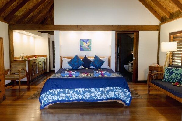 Un dormitorio con techo de madera, una cama con colcha azul estampada de flores, dos almohadas, una mesita de noche, una silla y un cuadro colgado en la pared.