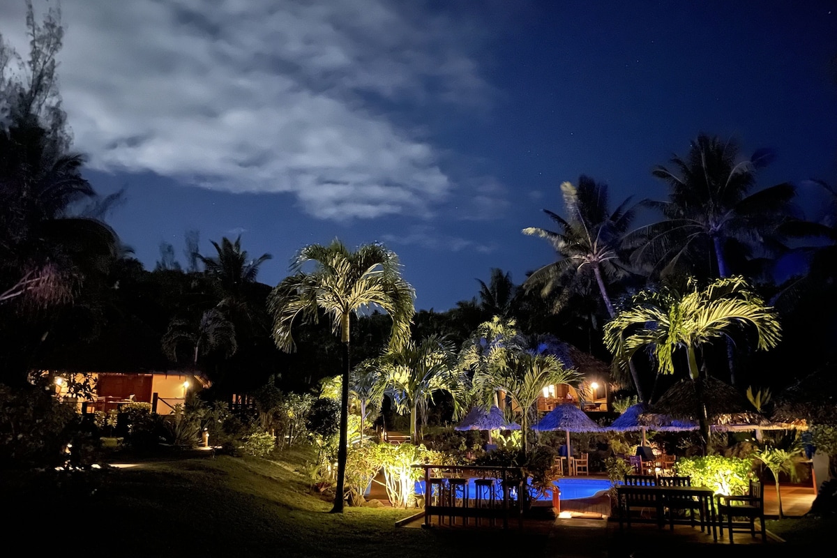 A nighttime resort with a pool, palm trees, and outdoor seating areas.