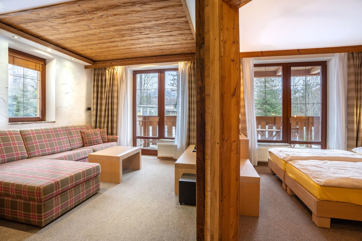 Een knus ingerichte kamer met een houten plafond, een geruite bank, een bed, een bureau en grote ramen met gordijnen.