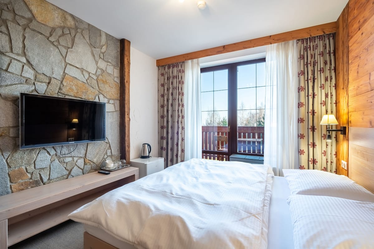 Slaapkamer met een stenen muur, houten balken, een groot bed, een televisie, een nachtkastje en een raam met gordijnen.