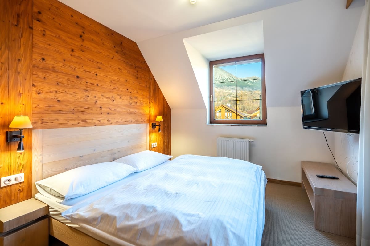 Een slaapkamer met een houten hoofdbord, een bed met witte lakens, een nachtkastje met een lamp, een televisie op een dressoir en een raam met uitzicht op de bergen.