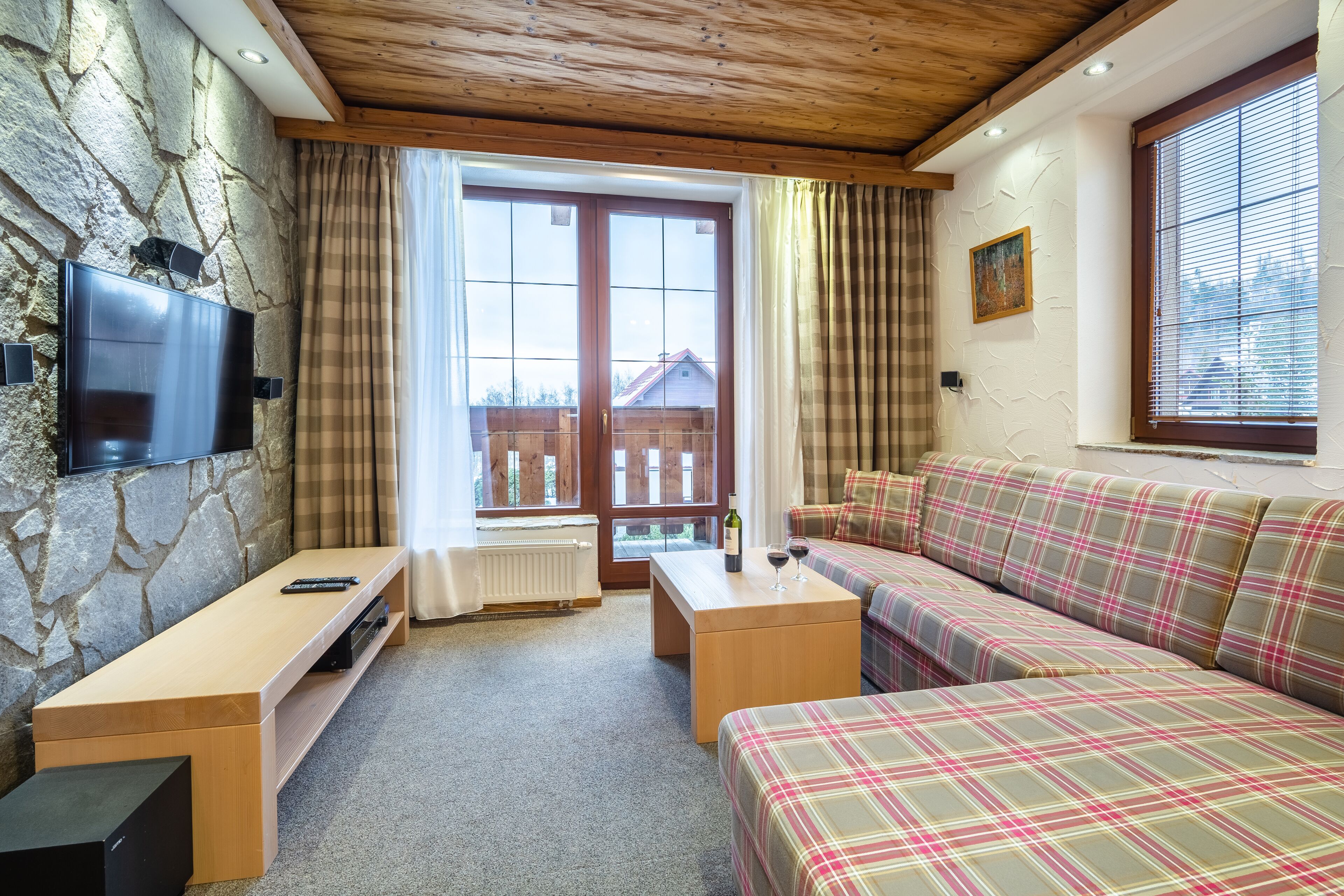 A cozy room with a stone wall, wooden ceiling, a flat-screen TV, a wooden table, a sofa, and a window with curtains.