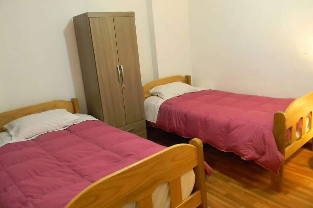 Una habitación con dos camas de madera, cada una con una colcha rosa y almohadas blancas. Hay un armario de madera de dos puertas y una silla de madera.