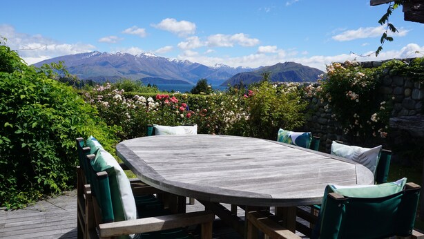 Um deck de madeira com uma mesa redonda e cadeiras, rodeado por vegetação exuberante e uma parede de pedra.