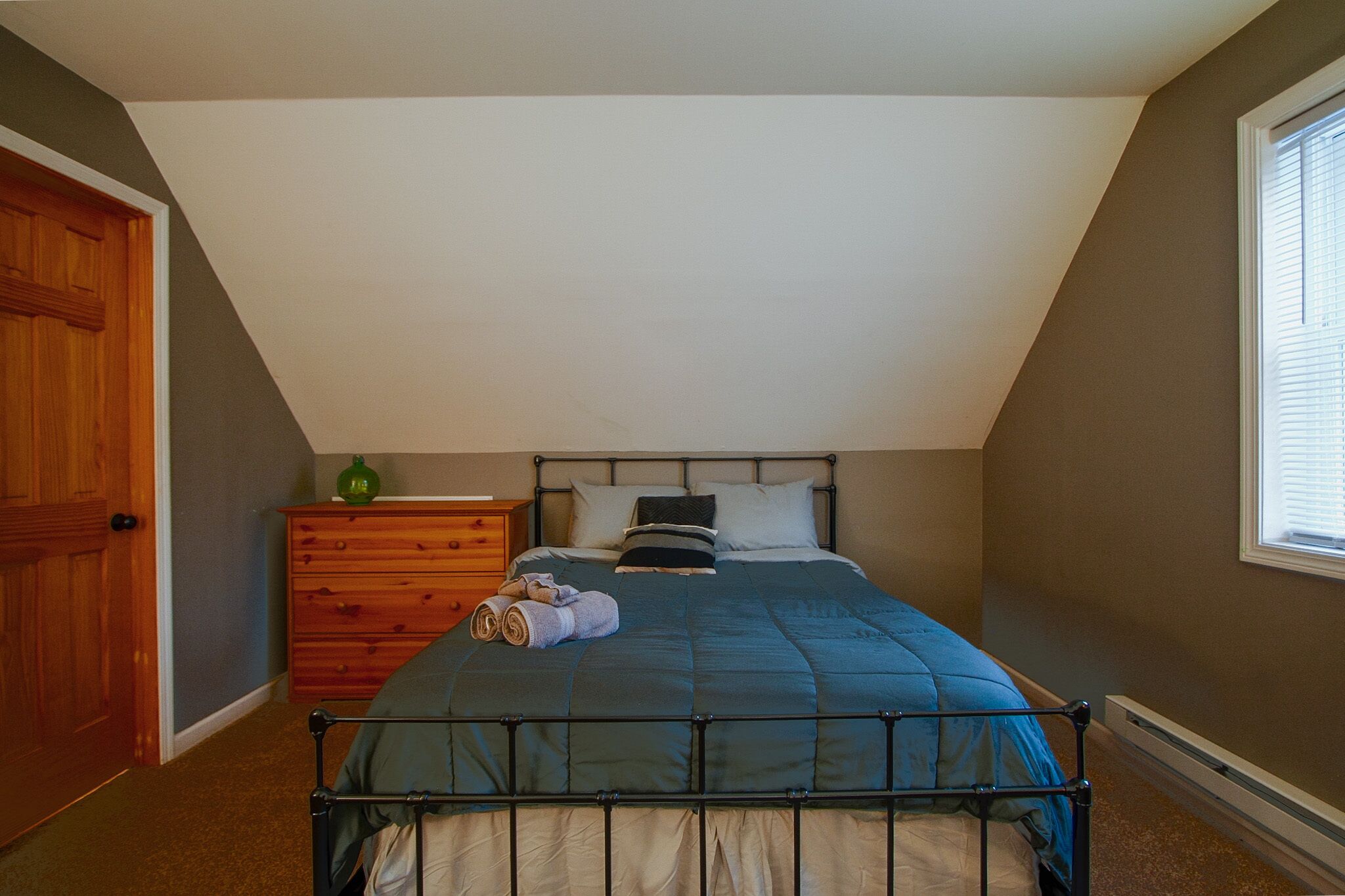 A bedroom with a sloped ceiling, a bed with a metal frame, a wooden dresser, and a window with blinds.