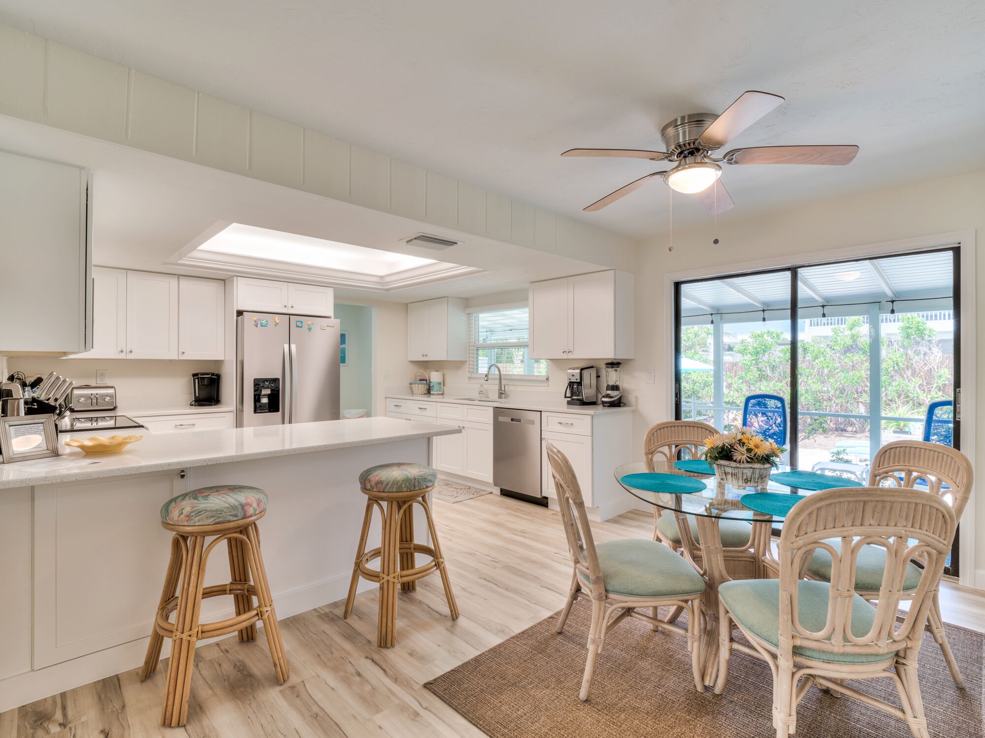 A modern kitchen with white cabinetry, a central island with stools, a dining area with wicker chairs, and a ceiling fan.
