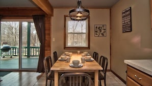 A dining area with a wooden table set for six, a hanging light fixture, and a window with blinds.