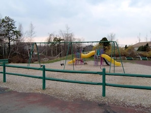 Un parc infantil amb cadira d'oscil·lació i tobogans, envoltat d'arbres i una zona de grava.