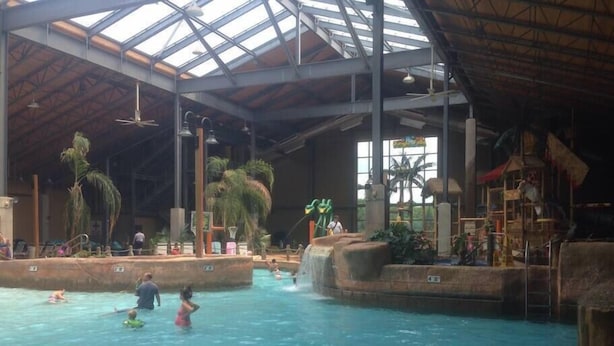 An indoor swimming pool with multiple lanes, palm trees, and a play area with slides and climbing structures.