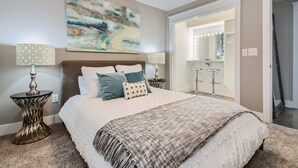 Ein Schlafzimmer mit einem großen Bett, zwei Nachttischen mit Lampen, einem Deckenventilator, einem großen abstrakten Gemälde und einem durch eine offene Tür einsehbaren Badezimmer.