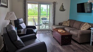Ein Wohnzimmer mit einer Couch, einem Sessel, einem Couchtisch, einer Lampe und einem Fernseher. An der Wand ist eine Stern-Dekoration angebracht und durch eine Schiebetür ist ein Blick ins Freie möglich.