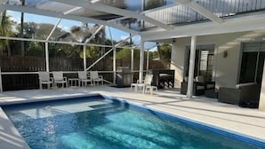 Una piscina con un patio cubierto, mobiliario exterior y una cerca de madera al fondo.