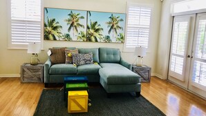 Una sala de estar con un sofá modular, dos mesitas auxiliares, un ventilador de techo y un cuadro de gran tamaño con temática tropical.