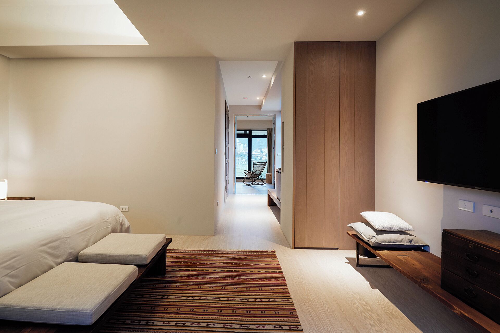 A modern hotel room with a large bed, a bench, a TV, and a view of the city through the window.