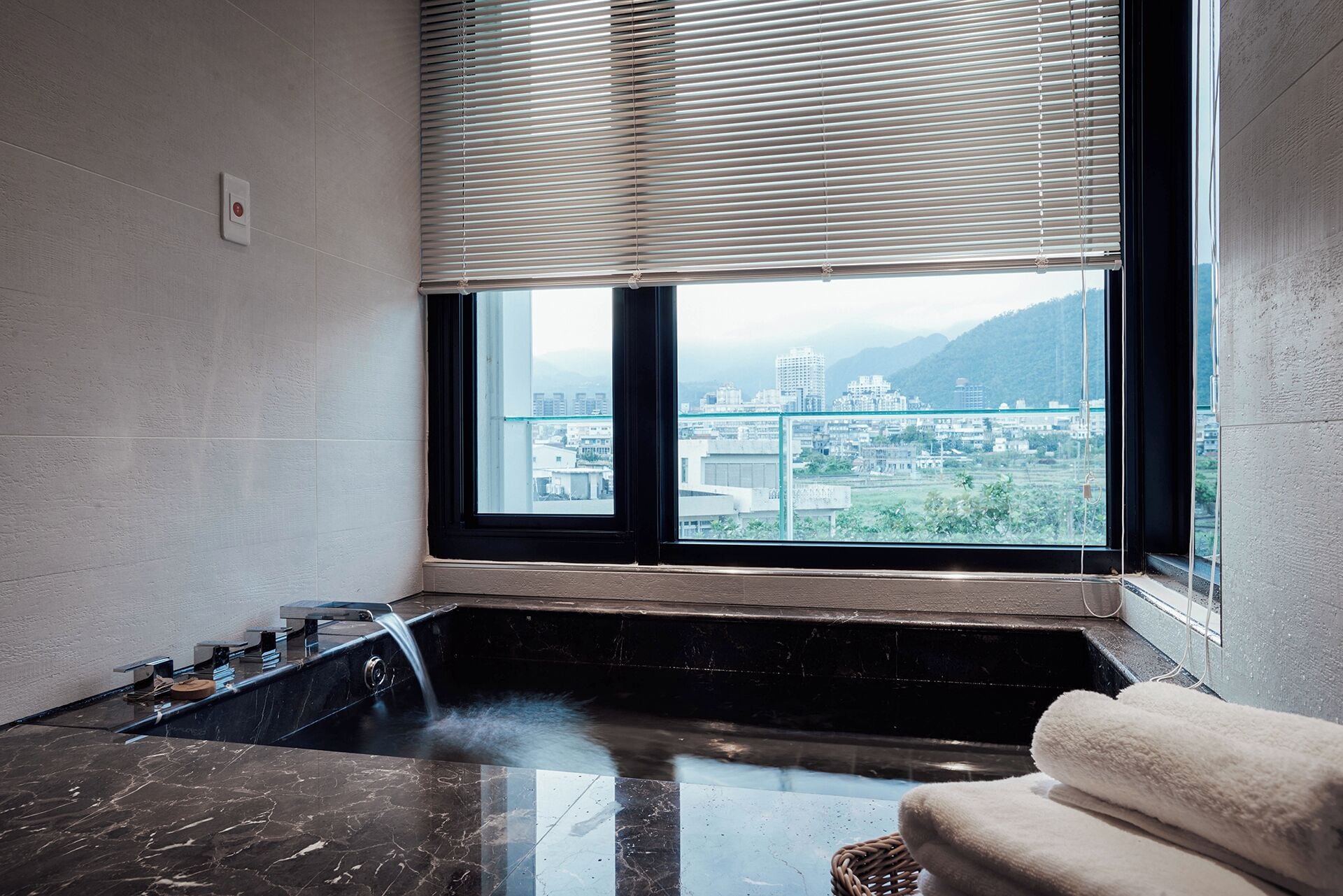 A bathroom with a bathtub, a window with blinds, and a view of a cityscape.
