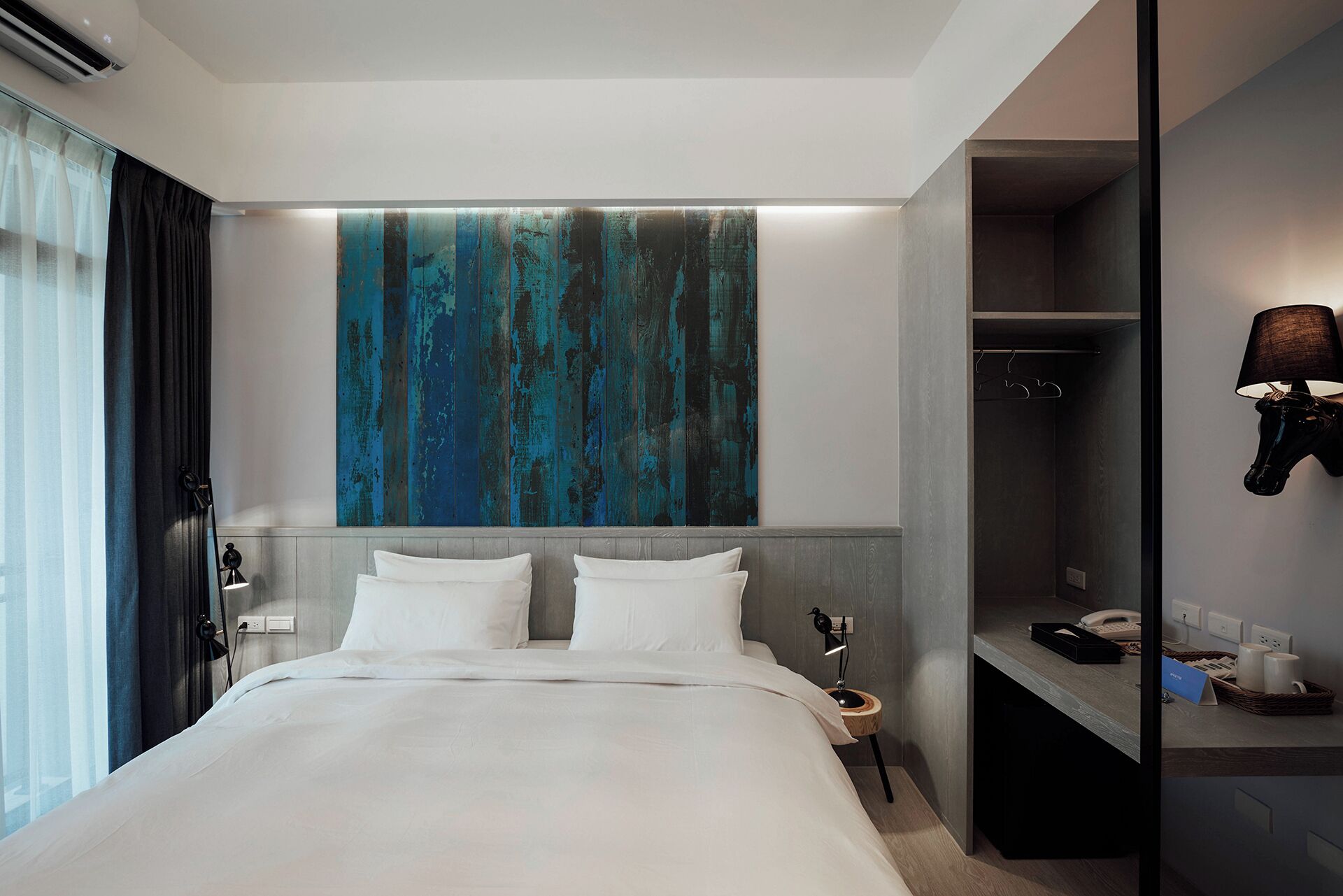 Une chambre d’hôtel moderne avec un grand lit, une armoire, un bureau avec une lampe et un grand tableau abstrait accroché au mur.