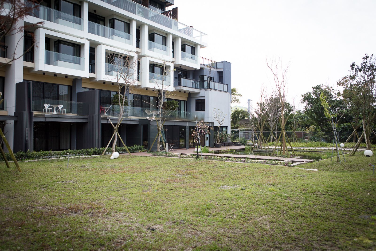 A modern multi-story building with balconies, a small garden area with trees and a pond, and a pathway leading to the entrance.