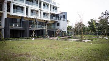 A modern multi-story building with balconies, a small garden area with trees and a pond, and a pathway leading to the entrance.