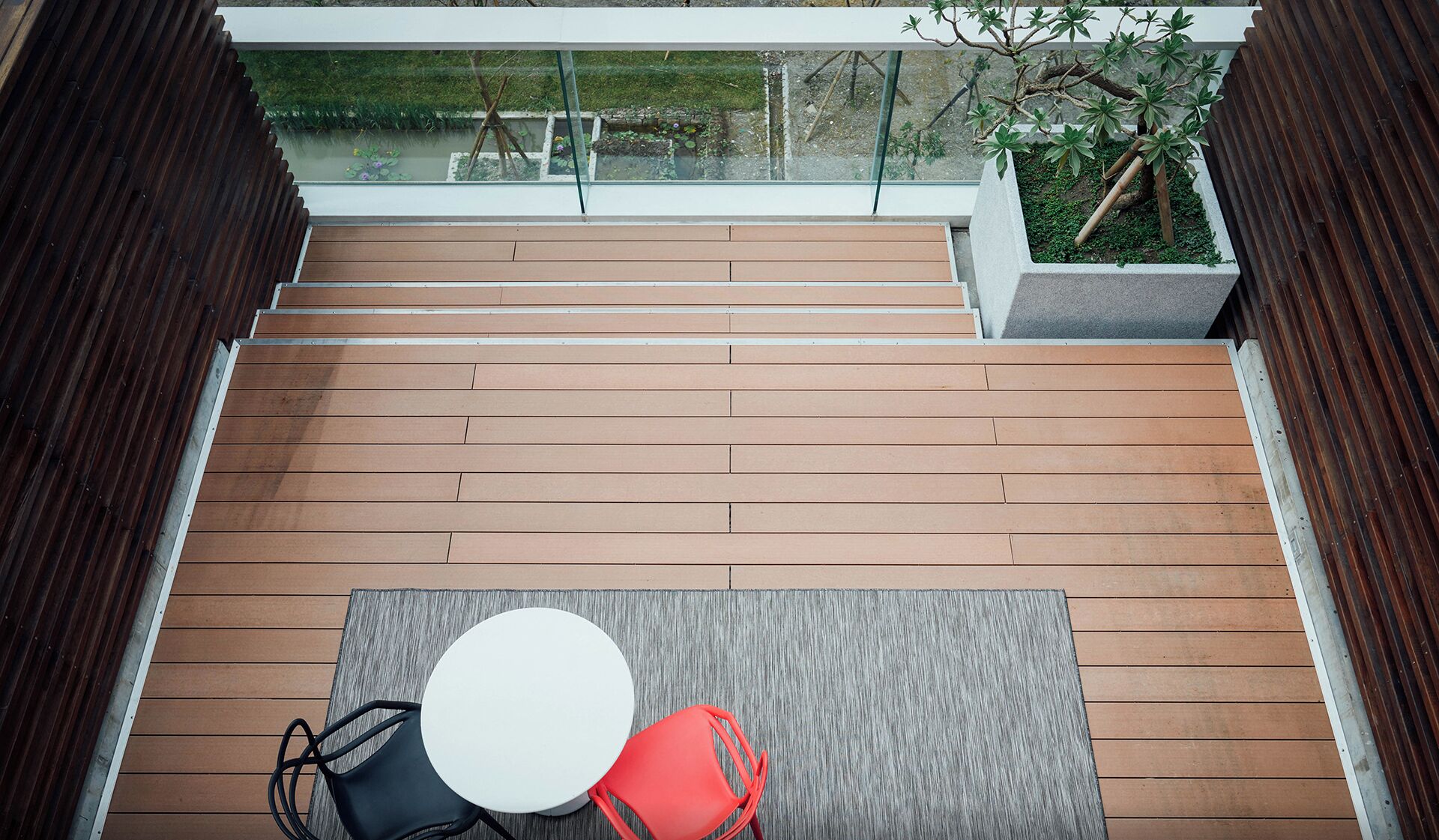 Een houten terras met een tafel, stoelen en een glazen balustrade, met uitzicht op een tuin.
