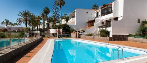 A swimming pool with a tiled deck, surrounded by white buildings with balconies, palm trees, and a clear blue sky.